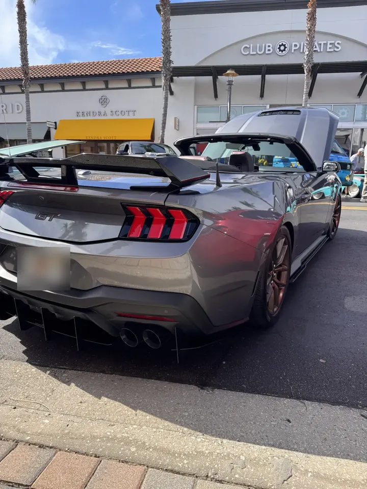 Gray 2026 Mustang GT convertible from the rear three-quarter, top down, with bronze wheels and a gloss black spoiler